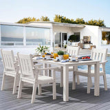 Outdoor dining area with white furniture on a wooden deck, featuring a house and ocean view.