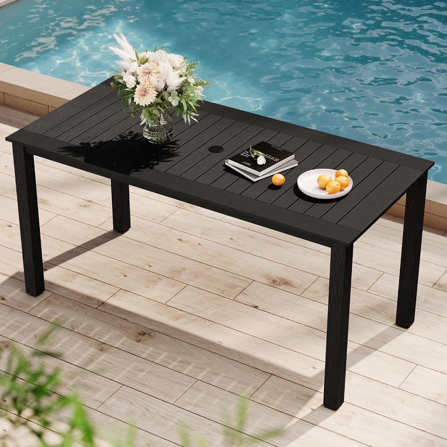 Black outdoor table with a vase of flowers, books, and a plate of oranges by a pool.